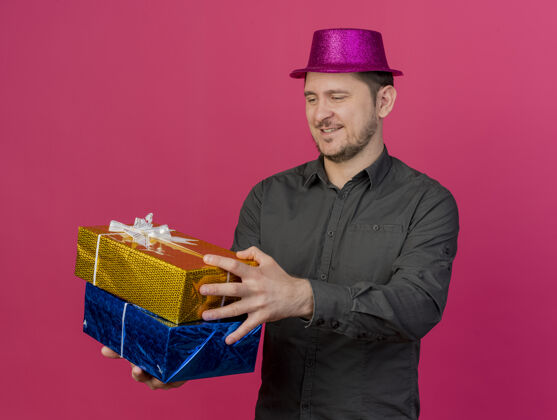 礼物高兴的年轻人戴着粉红色的聚会帽 拿着礼品盒在一旁孤立的粉红色壁板派对年轻人