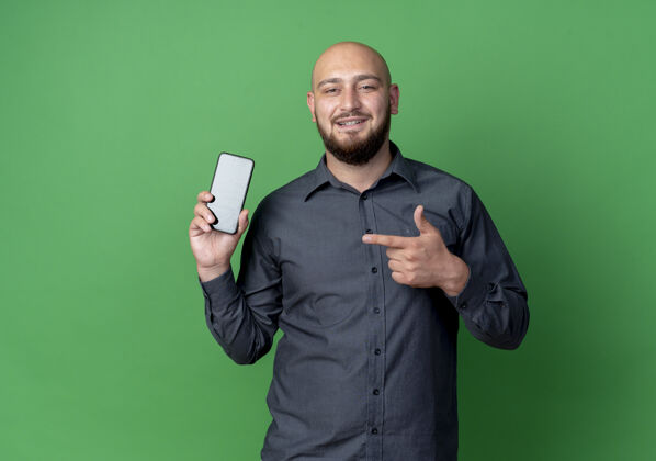 感觉微笑的年轻秃头呼叫中心男子显示和指着手机隔离在绿色的墙壁上衣服电话姿势