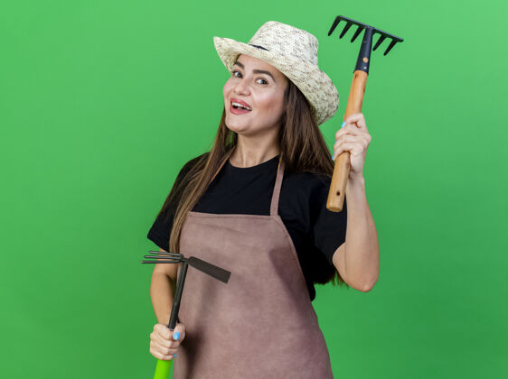 帽子快乐美丽的园丁女孩穿着制服戴着园艺帽手持锄头耙和提高耙隔离在绿色提高耙举行