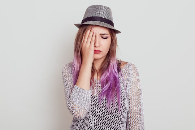 痛苦心烦意乱的美丽女人 淡紫色头发 穿着衬衫 戴着帽子 悲伤 用手掌遮住半张脸 闭着眼睛 隔着白墙困难坏情绪压力