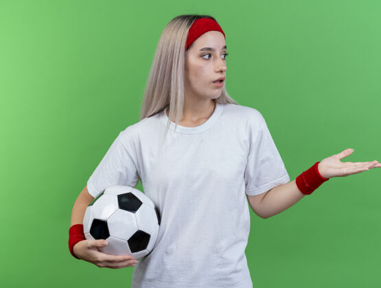 女孩令人印象深刻的年轻运动女性戴着背带 戴着头带和腕带 拿着球看着并指着隔离在绿色墙上的一侧印象腕带女人