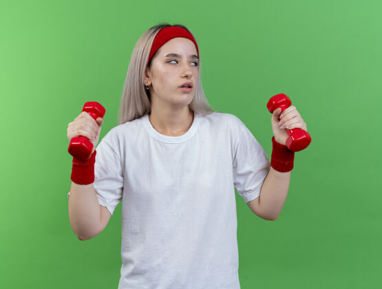 穿恼怒的年轻运动女子戴着背带 戴着头带和腕带 眼睛转动 举着哑铃孤立地站在绿色的墙上腕带人女人