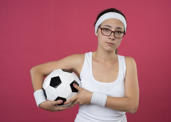 光学戴着眼镜 戴着头带和腕带的失望的年轻运动女性把球孤立地举在粉红色的墙上球人腕带