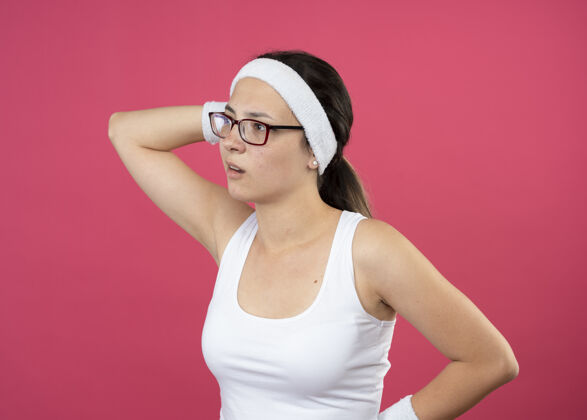 头笨手笨脚的年轻运动女人戴着眼镜 戴着头带和腕带 抱着头 看着粉色墙上孤立的一面人年轻眼镜