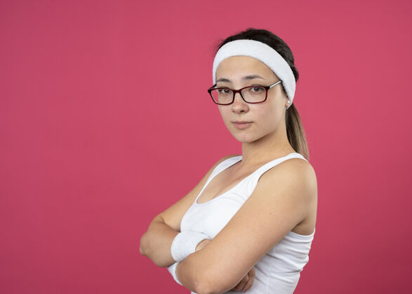 手臂自信的年轻运动女性戴着眼镜戴着头带和腕带站在一边 交叉双臂孤立在粉红色的墙上腕带穿年轻