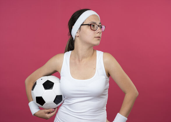 人自信的年轻运动女性戴着眼镜戴着头带和腕带拿着球 看着粉色墙上孤立的一面腕带头带运动