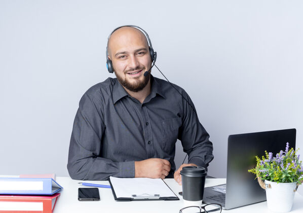 工具微笑的年轻秃头呼叫中心男子戴着耳机坐在办公桌旁 手放在办公桌上 隔离在白色的墙上脸穿着人