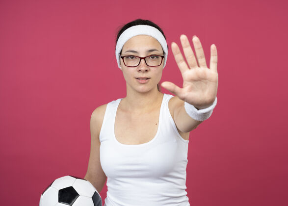 女人自信的年轻运动女性戴着眼镜戴着头带和腕带拿着球 手势停止手势孤立在粉红色的墙上自信手势眼镜