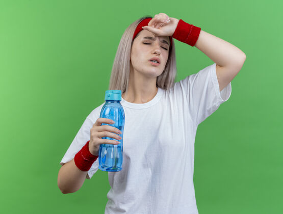 人带着护腕戴着头带和腕带的疼痛的年轻运动女性把手放在额头上 把水瓶隔离在绿色的墙上戴头带抱着