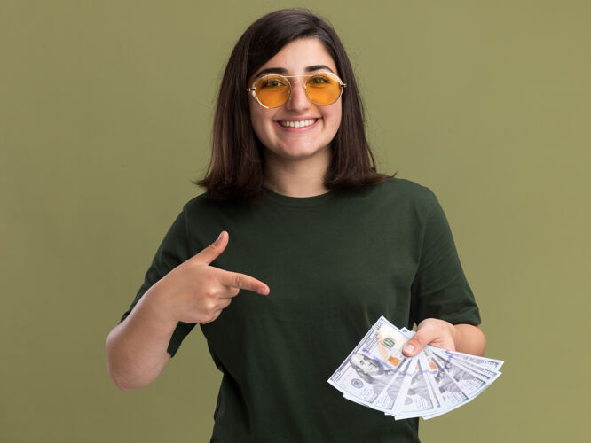 钱微笑着的年轻漂亮的高加索女孩 戴着太阳眼镜 拿着橄榄绿的钞票 指着钞票绿漂亮微笑