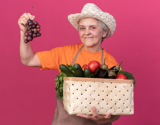 园艺高兴的上了年纪的女园丁戴着园艺帽 手里拿着菜篮子和一串粉红色的葡萄束女花园