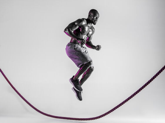 人世界之上以灰色工作室为背景的年轻的非洲裔美国健美运动员训练穿着运动服的肌肉男模特跳过战绳运动的概念 健美 健康的生活方式运动鞋健美力量