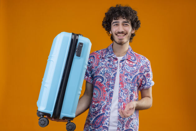 旅行者快乐的年轻帅气的卷发旅行者在孤立的橙色空间里拿着手提箱男人帅气手提箱