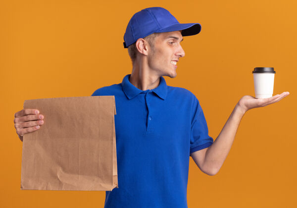 包装微笑的年轻金发送货男孩拿着纸包裹 看着橙色的杯子送货微笑年轻