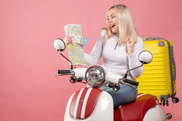 漂亮前视图快乐的金发女孩骑着轻便摩托车拿着票和地图轻便摩托车坐着前面