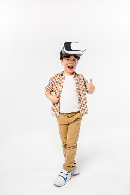 科学快乐和好奇穿着牛仔裤和衬衫 戴着虚拟现实耳机眼镜的小男孩或孩子 背景是白色的工作室尖端技术的概念 视频游戏 创新虚拟现实视觉耳机