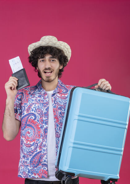 快乐快乐的年轻英俊的卷发旅行者戴着帽子拿着钱包和机票和手提箱在孤立的粉红色空间年轻人穿着手持