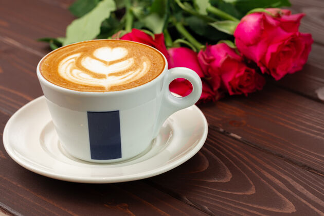 浪漫木桌上放着一杯卡布西诺咖啡和一束玫瑰礼物花瓣庆祝