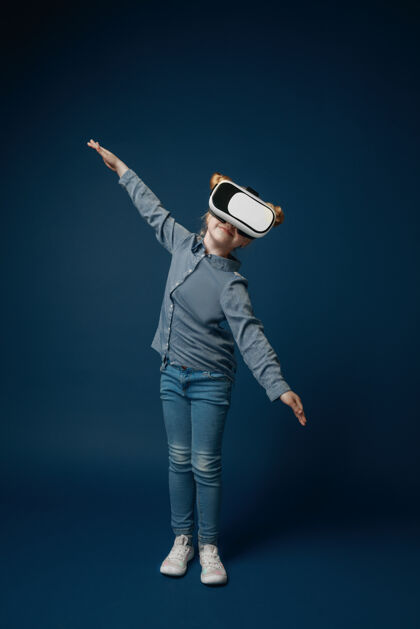 科技像飞机一样飞行穿着牛仔裤和衬衫的小女孩或孩子 蓝色工作室背景上的虚拟现实耳机眼镜尖端技术的概念 视频游戏 创新创新眼镜未来