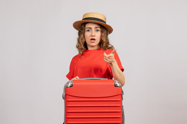 旅行箱假日女孩的正面图 她的手提箱指向前面站在白色的墙上包快乐漂亮