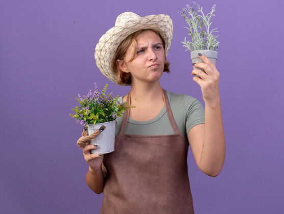 年轻体贴的年轻斯拉夫女园丁戴着园艺帽捧着紫色花盆里的花看园艺斯拉夫花园