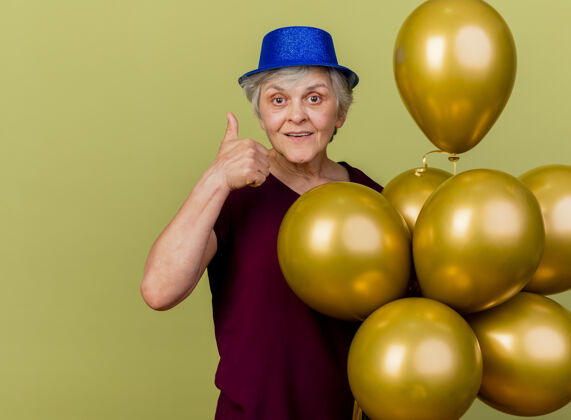 帽子戴着派对帽的快乐老妇人站在橄榄绿的墙上 站着氦气球 竖起大拇指 留着复制空间氦请橄榄