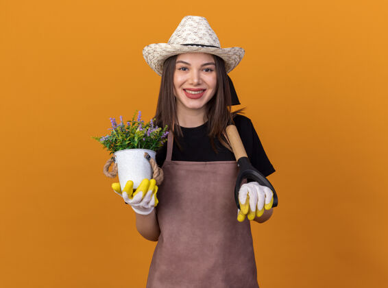 花盆微笑着美丽的白人女园丁戴着园艺帽和手套 肩上扛着花盆和铁锹放在橘子上园艺黑桃肩膀