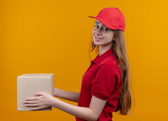 观看微笑着的年轻送货女孩 穿着红色制服 站在孤立的橙色空间的侧视图中侧面制服橙色