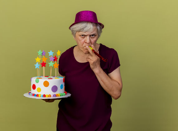 帽子自信的老妇人戴着聚会帽吹着口哨拿着生日蛋糕孤立地放在橄榄绿的墙上 还有复制空间生日老人橄榄