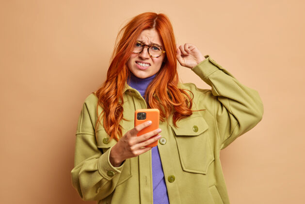 白种人不高兴的红发女子搔头皱眉从不满试图解决问题的智能手机不知道如何使用新的应用程序穿着时尚的衣服问题电话姿势