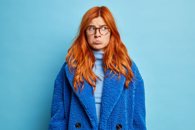 眼镜年轻的不高兴的女人把嘴唇包起来 表情阴沉地站在一边 穿着高领毛衣和蓝色冬衣担心人女人