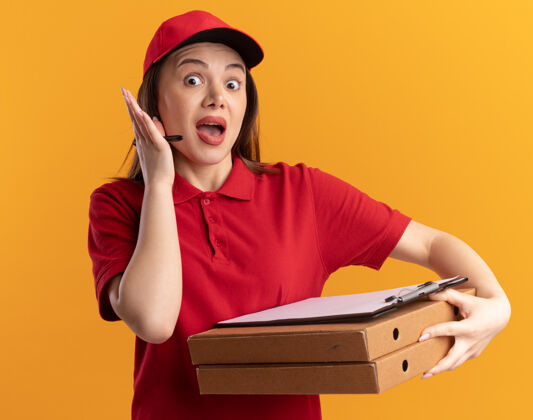 漂亮穿着制服的焦急漂亮的女送货员拿着马克笔和剪贴板放在橘色的比萨饼盒上焦虑剪贴板记号笔