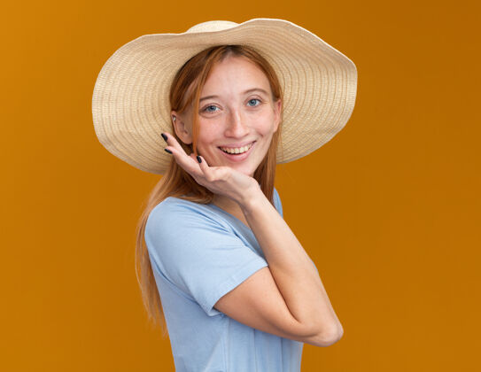 帽子带着雀斑的年轻红发姜黄色女孩微笑着 戴着沙滩帽 站在一旁 手放在橙色的下巴上微笑姜沙滩