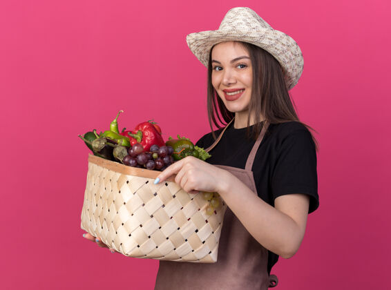 蔬菜笑容可掬的白人女园丁戴着园艺帽捧着菜篮子看着粉色的相机花园女园艺