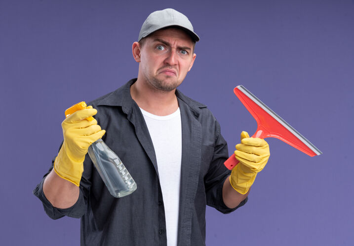 不愉快年轻帅气的清洁工穿着t恤 戴着帽子和手套 拿着拖把头和喷雾瓶隔离在紫色的墙上人喷雾手套