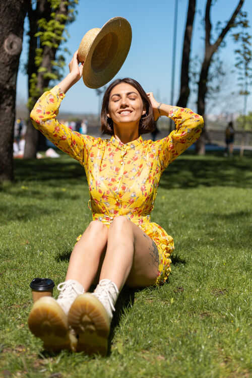 女人穿着黄色夏装戴着帽子坐在公园草地上的女人的户外肖像桥梁阳光自然