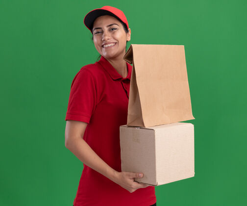 食物微笑的年轻送货女孩穿着制服 戴着帽子 拿着一个纸食品包装盒 隔离在绿色的墙上脸年轻帽子