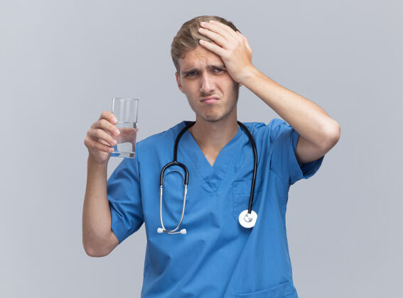 听诊器遗憾的是 年轻的男医生穿着医生制服 手持听诊器 把一杯水放在隔离在白墙上的头上后悔头玻璃