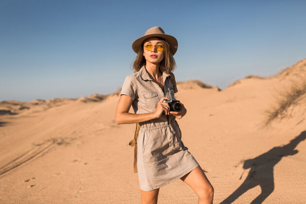 时尚穿着卡其色衣服的时髦年轻女子在沙漠中行走 在非洲旅行 戴着帽子和背包 用老式相机拍照自然旅行者相机