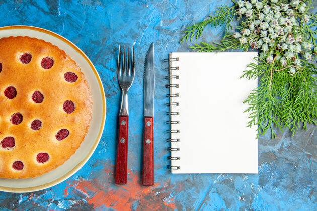 工具树莓蛋糕的俯视图在椭圆形的圆锥形盘子上叉子刀在蓝色的表面上一个笔记本刀艺术家钢笔