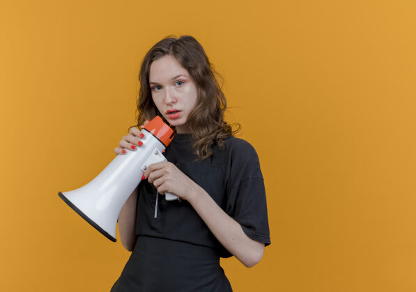 女性年轻的斯拉夫女理发师穿着制服 拿着扬声器 看着隔离在橙色背景上的相机 还有复印空间背景发言人年轻