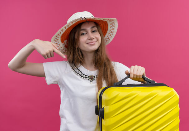 穿自信的年轻旅行家女孩戴着帽子拿着手提箱 指着它在孤立的粉红色空间手提箱女孩自信