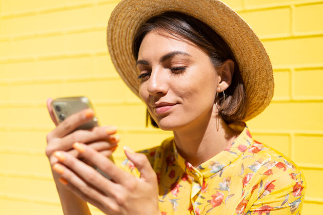可爱穿着黄色夏装戴着黄色帽子的女人在黄色的砖墙上冷静而积极地拿着手机休闲显示脸