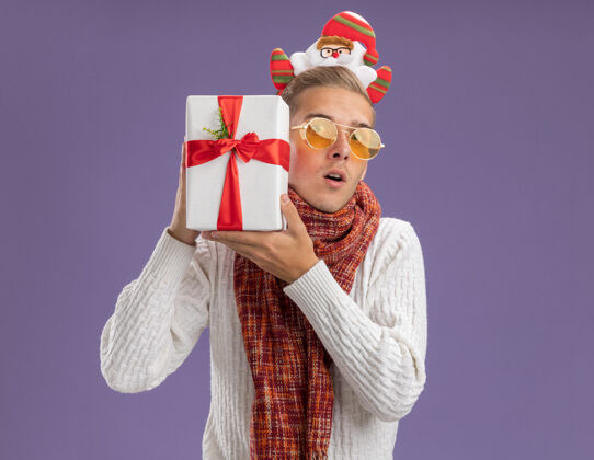 圣诞老人好奇的年轻帅哥戴着圣诞老人的头带和围巾拿着礼包靠近头部看着隔离在紫色背景上的相机圣诞快乐头带围巾