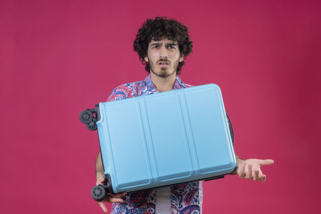 拿着困惑的年轻英俊的卷发旅行者男子拿着手提箱 在孤立的粉色空间和复印空间上展示空手年轻困惑手提箱