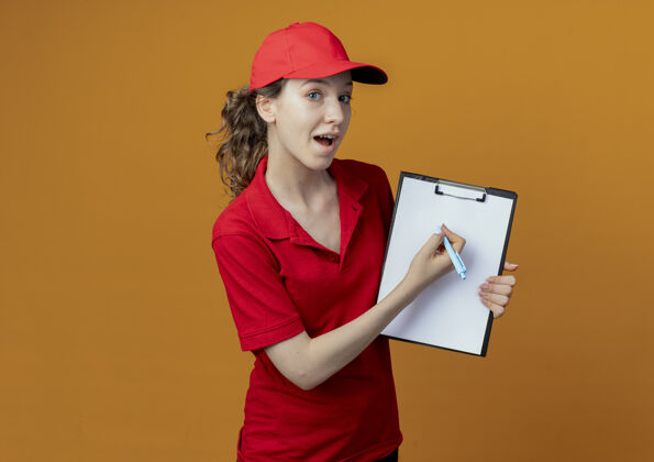 年轻令人印象深刻的年轻漂亮的送货女孩穿着红色制服 戴着帽子 拿着钢笔和剪贴板 看着隔离在橙色背景和复印空间的相机交货印象举行