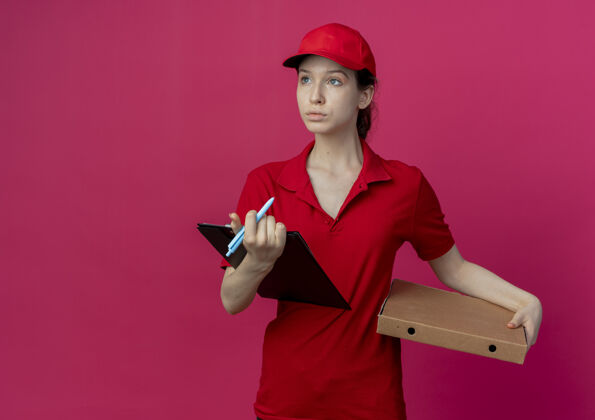 包装年轻漂亮的送货女孩 穿着红色制服 戴着披萨包装笔帽 拿着剪贴板 看着被隔离在深红色背景上的一面 还有复印空间钢笔漂亮年轻
