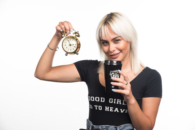 姿势在白墙上拿着钟和咖啡杯的年轻女人金发杯子时钟