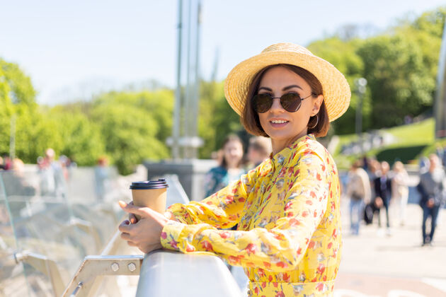 年轻人穿着黄色夏装 戴着黄色帽子 喝着咖啡 享受阳光的女人的户外肖像 站在桥上 城市的景色令人惊叹欢乐城市度假