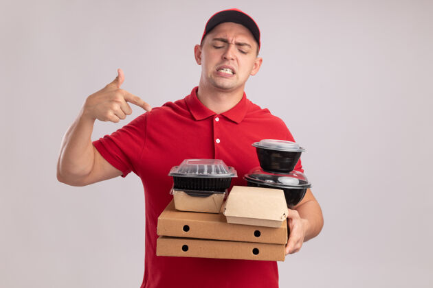 点年轻的送货员穿着制服 戴着帽子 指着隔离在白墙上的比萨饼盒上的食品容器感觉制服人